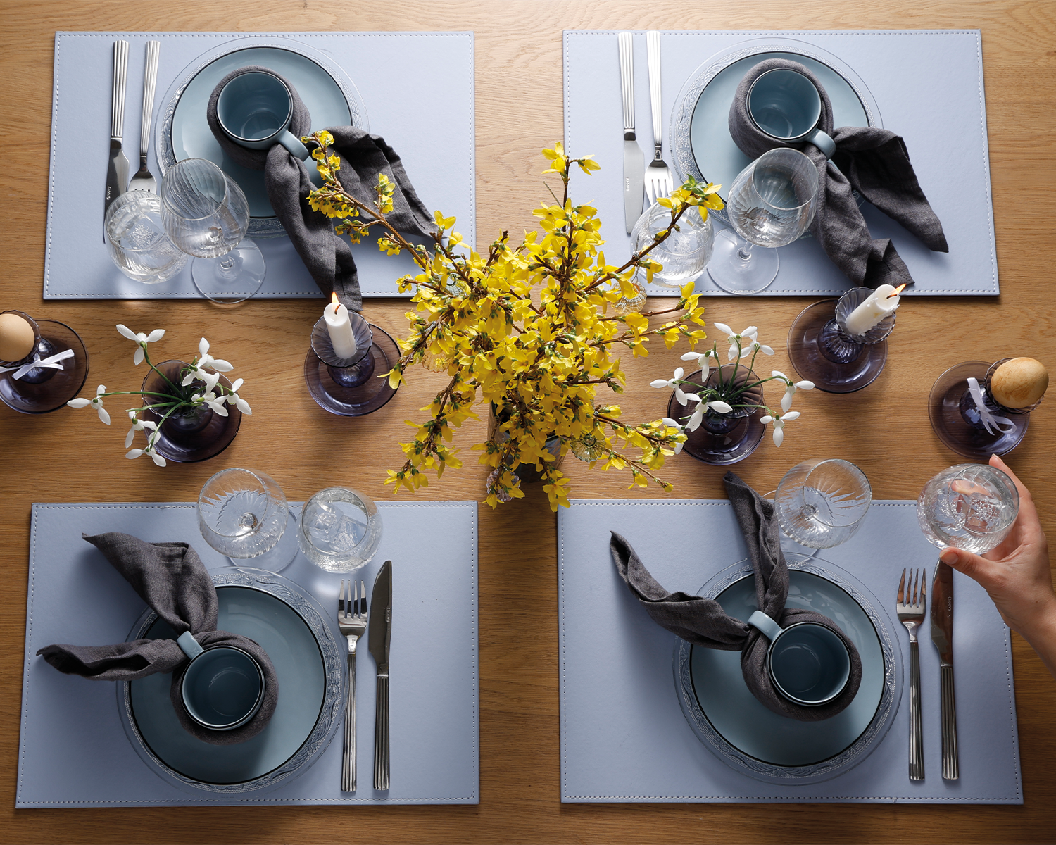 Påskebord dækket med blå dækkeservietter, blåt stel, og stofservietter. På midten af bordet står der blå stager med lys, blomster og æg. I midten står en forårsbuket med gule blomster. En arm rækker ind og stiller et vandglas på bordet.