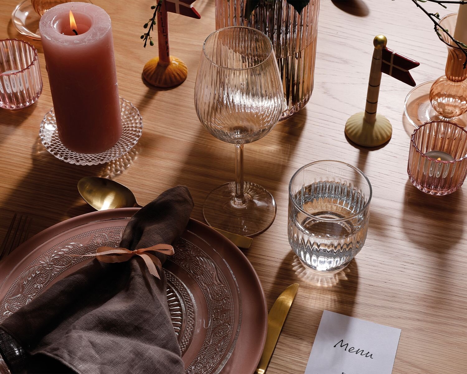 Træbord dækket med lyserød og glas tallerken. Klare vinglas og vandglas med mønster. Guld bestik og menu til gæsterne.