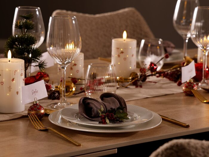 Et smukt opdækket julebord med hvide tallerkener, gyldent bestik, krystalglas og tændte stearinlys med julemotiver. Bordet er pyntet med røde julekugler, grønne grene og navnekort, der skaber en varm og festlig julestemning.
