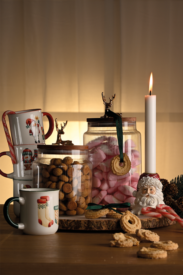 Stemningsfuldt julebord med julekrus med motiver, glaskrukker med pebernødder og juleskum, omringet af vaniljekranse, gran og en julemandslysestage.