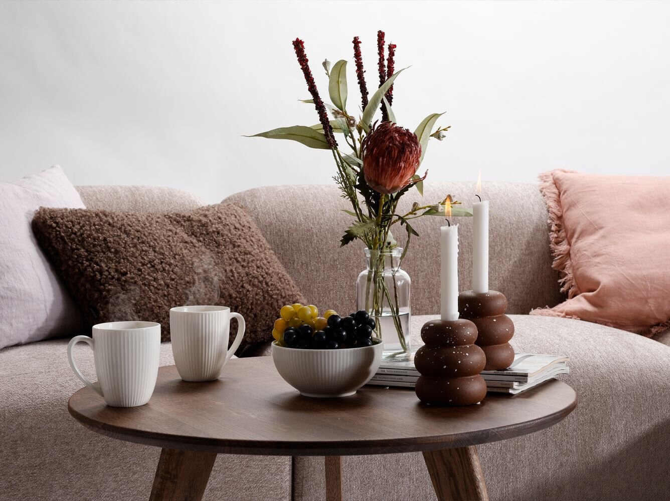 Mørkt rundt træsofabord med lysestager, blomster i vase, en skål med vindruer og to kopper kaffe. Bag bordet kan man se en beige sofa med puder i brun, pastel lilla og en rosa pude med frynser.