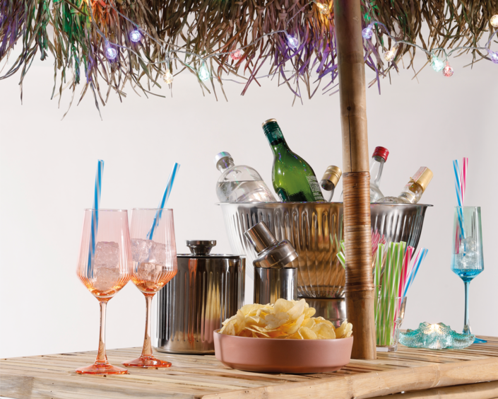 Udendørs baropsætning med farverige glas, iskolde flasker, drinks og lyskæde under stråtag skaber en festlig sommerstemning.