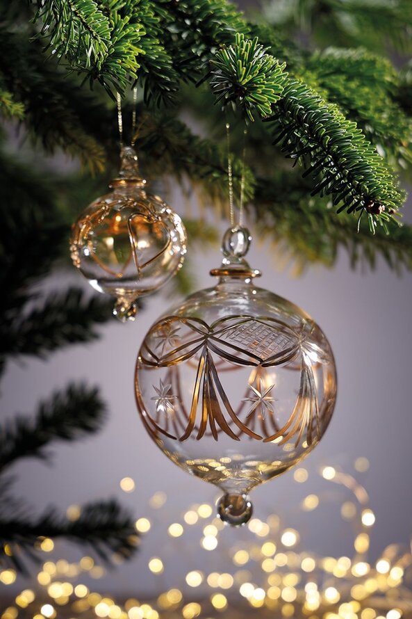 Glas julekugler i to størrelser med gulddetaljer hænger fra grangrene med glitrende lys i baggrunden.