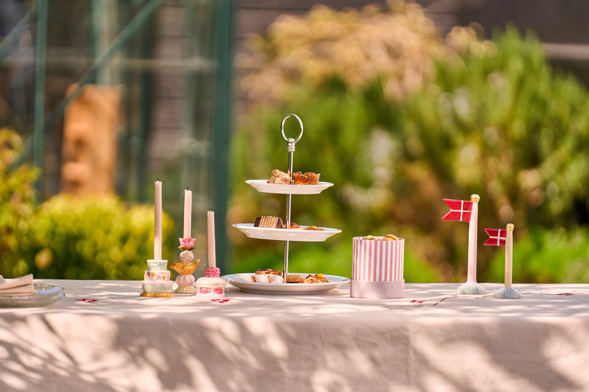 Havefest i eftermiddagsolen med blomster, lyskæder i træerne, og gæster der sidder rundt om borde i haven.