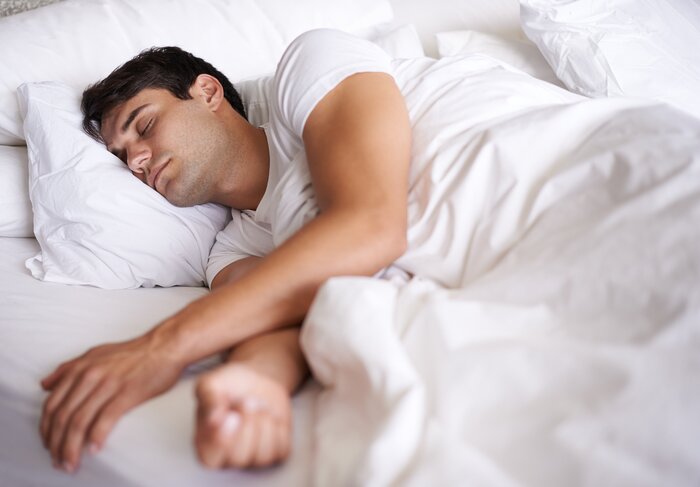 Mand sover på siden i en hvid seng med den ene arm hvilene mod kanten af sengen.
