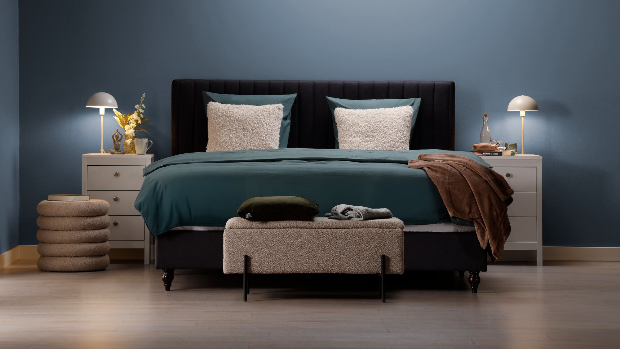 Soveværelse med blå væg, stor seng i midten med blåt sengetøj og beige pyntepuder. Soveværelset er også pyntet med to hvide sengeborde med 3 skuffer og en hvid bordlampe. Udover det er der en beige bænk for fodenden  af sengen, samt en rund beige puf.