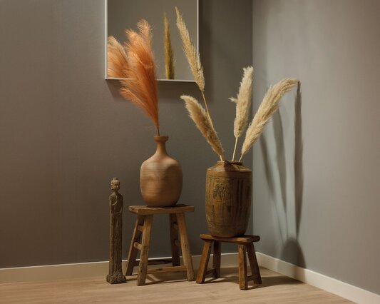 Hjørne af et rum indrettet med boho interiør, små rustikke træskamler med store keramik vaser fyldt med pampas græs. På gulvet ved siden af står en høj Buddha figur og over hænger et hvidt buet spejl.