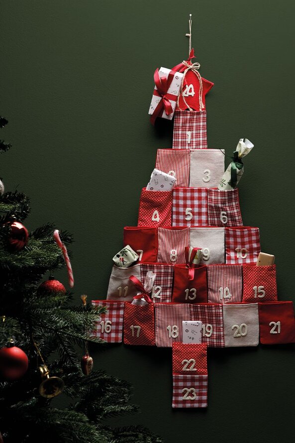 Julekalender formet som et juletræ med røde og hvide stofposer med tal, hængende på en væg ved siden af et pyntet juletræ.