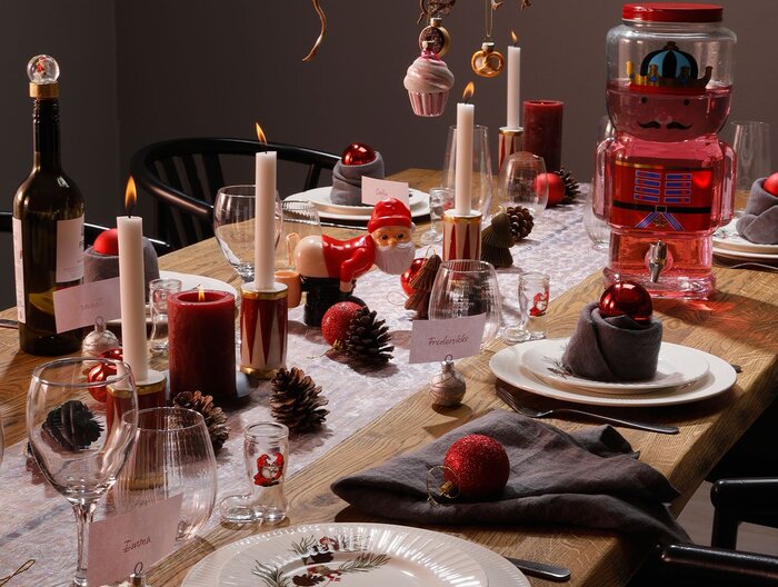 Et stemningsfuldt julebord pyntet med røde stearinlys, kogler, julekugler og tallerkener med grå servietter og glas. På bordet står også små julefigurer, herunder en nisse og en stor glaskande formet som en tinsoldat fyldt med rød drik. Navnekortene og det varme lys skaber en hyggelig og festlig atmosfære.