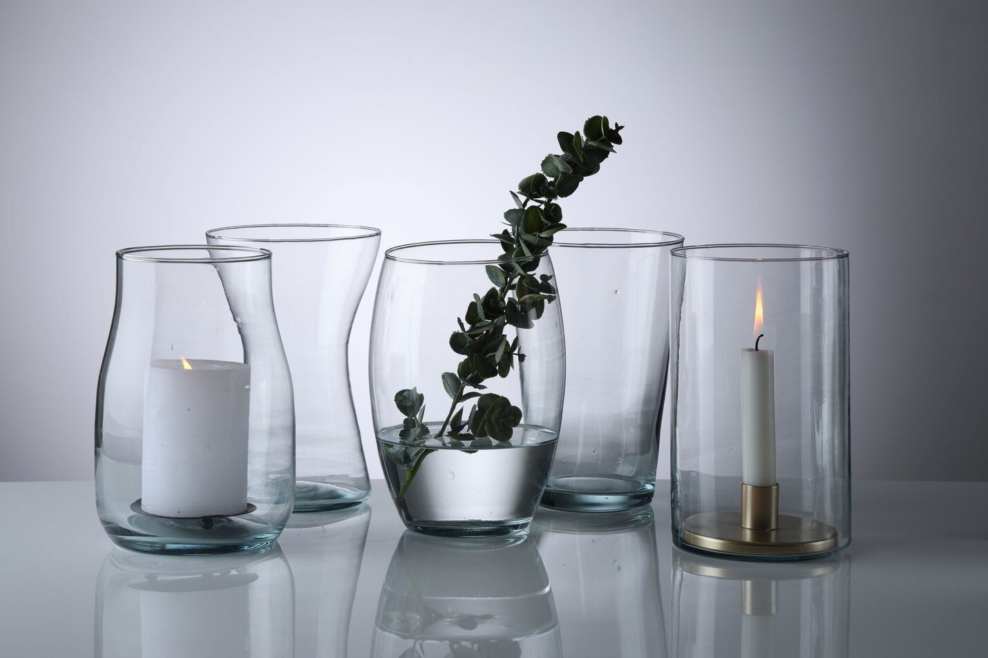 Fem klare glasvaser i forskellige former pyntet med eucalyptus gren, bloklys og stearinlys.