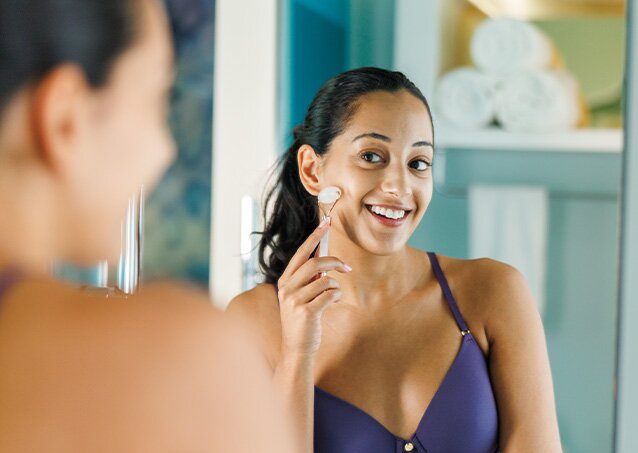 A woman smiles while looking in the mirror as she does her skincare routine in an image for a blog titled Why Does My Chin Hair Grow So Fast?