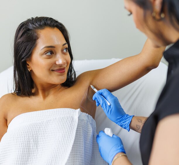 Woman raising her arm above her head and looking left as a Milan clinic staff member draws a grid on her underarm