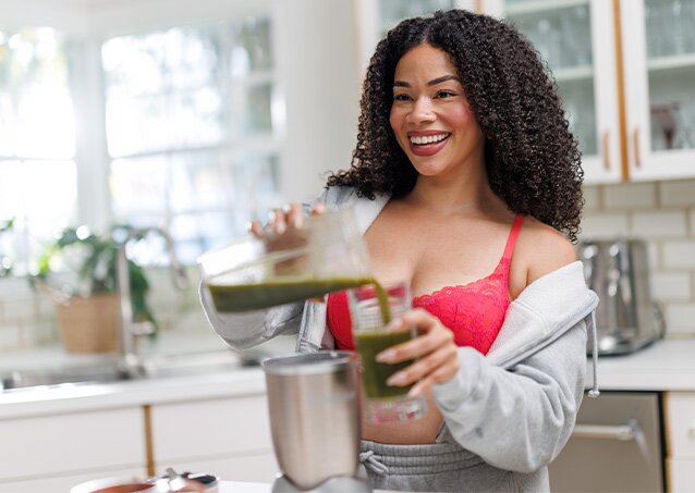 Happy woman in loungewear smiling and pouring a green smoothie into a glass
