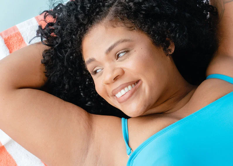 Woman wearing a blue bikini with curly hair holding her arms above her head after getting chin laser hair removal