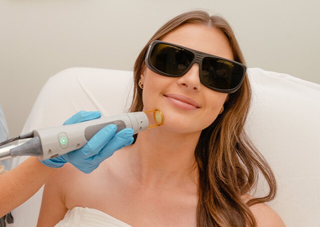 A woman wearing protective eyewear and a white towel gets laser hair removal on her chin in an image for a blog titled Laser Hair Removal vs. Plucking.