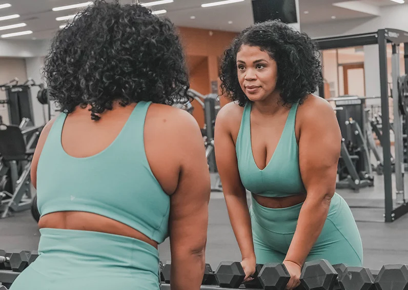 Woman in the gym in a green workout outfit grabbing weights and looking in the mirror