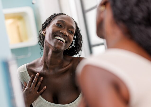 A woman wearing a white top smiles into the mirror because she has smooth skin without unwanted hair for a blog titled Why Does My Facial Hair Grow So Fast?