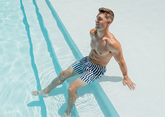 Muscular man with a trendy haircut in white and blue striped shorts smiling while dipping his legs in a pool