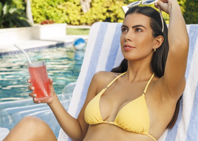 Gorgeous woman with black hair and a yellow bikini smiling and sitting in a pool chair while holding a drink