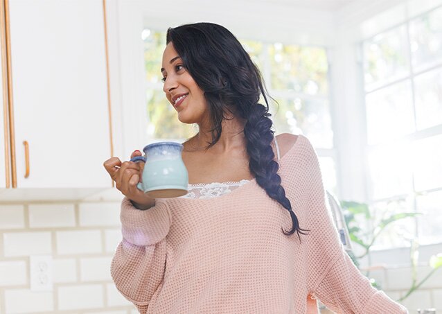 A woman wearing a pink sweater sips on some coffee in her kitchen in an image for a blog titled Laser Hair Removal On Your Period.