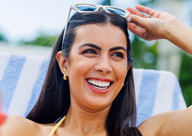 Woman with sunglasses on her head smiling and wearing a yellow swimsuit while sitting on a pool chair with a blue and white towel on it