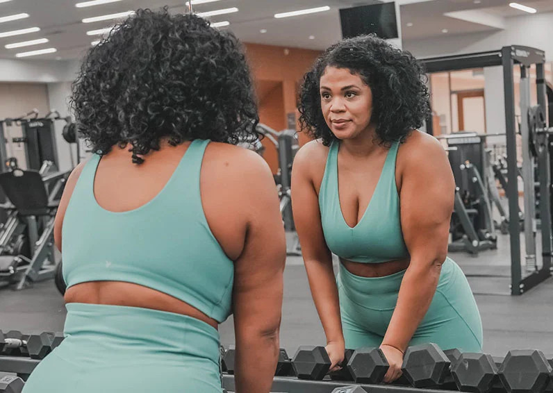 Woman in a teal athletic clothes set grabbing weights in a gym and looking in a mirror