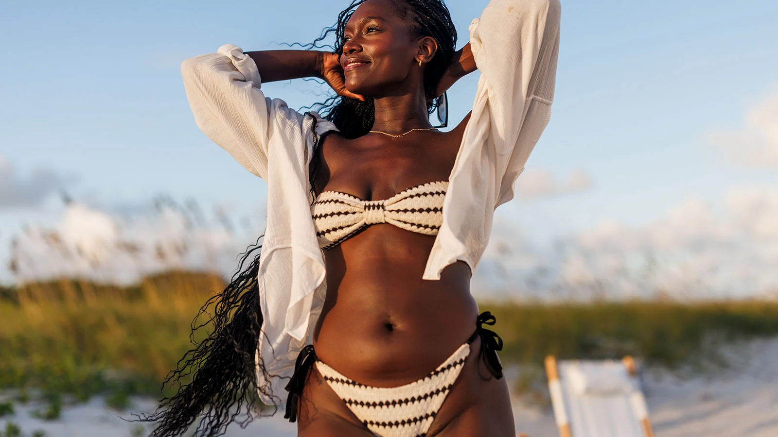 Woman in a bikini smiling on a beach while letting her white button-up shirt flow over her arms after getting treated with Brazilian laser hair removal at a Milan Laser clinic