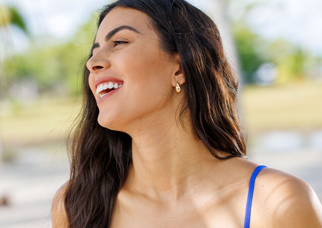 Woman with brunette hair and fair skin in a blue strappy shirt smiling in front of blurred vegetation