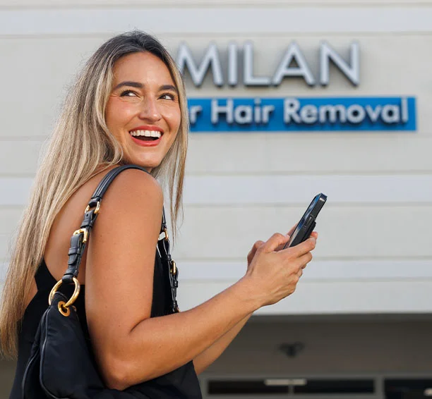 Smiling blonde woman holding a phone in front of a Milan Laser Hair Removal clinic, on her way to go get full-body laser hair removal treatments