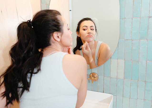 A woman wearing a light-blue top looks in the mirror for a hair to tweeze as she gets ready for the day in an image for a blog titled Laser Hair Removal vs. Tweezing.