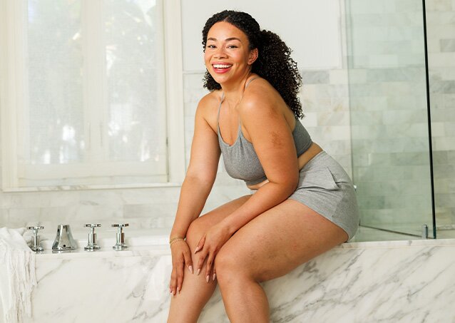 Woman in a light green sports bra and grey shorts sitting on a bathtub ledge and smiling