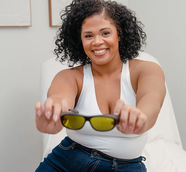 Woman holding up glasses and smiling while sitting in a treatment char during her consultation for laser hair removal for pilonidal cysts