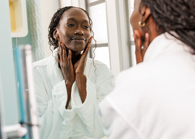 A woman wearing a white robe looks in the mirror while doing a skincare routine in an image for a blog titled Laser Hair Removal on Scars.