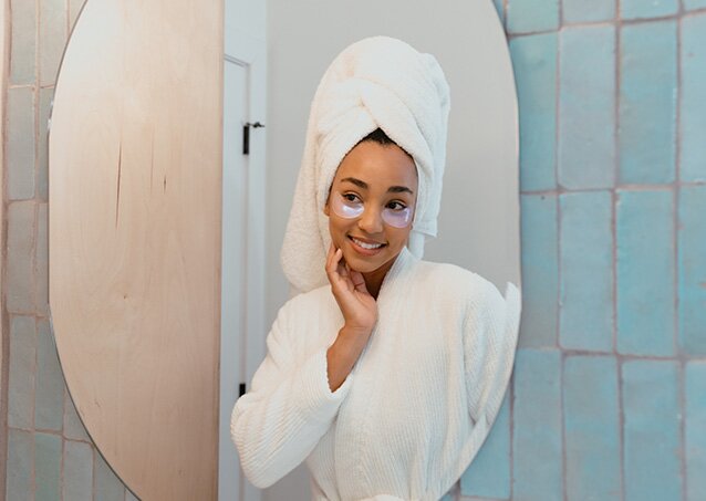 A woman wearing a white robe with a white towel wrapped around her head looks in the mirror and does her skincare routine for a blog titled Laser Hair Removal and Hair Growth Cycles.