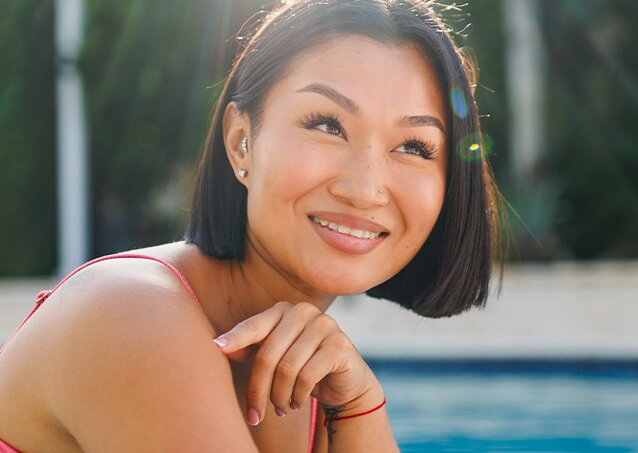 A woman in a pink bikini smiles while sitting next to the pool and enjoying the sun because she just learned How To Get Rid Of A Unibrow. 