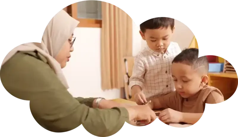 Seorang pengasuh sedang membacakan buku cerita kepada dua balita yang tersenyum di dalam ruang kelas daycare.