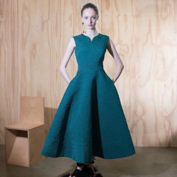 A model wearing a teal fit & flare midi dress made from textured cloqué, featuring a notched neckline with a signature sculpturally cut back.
