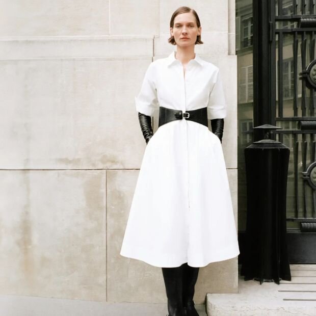 A woman wearing a white button-up A-line midi dress with a black leather belt.