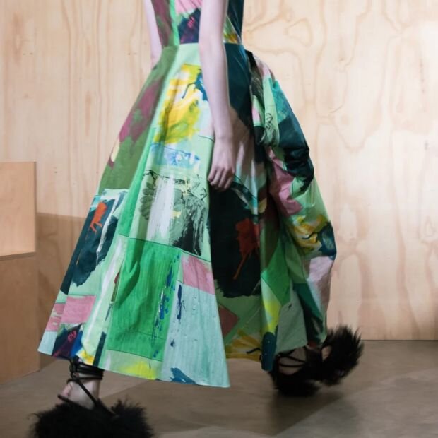 A model wearing an abstact multicolor pleated midi dress, featuring an asymmetrically draped bow at the waist.