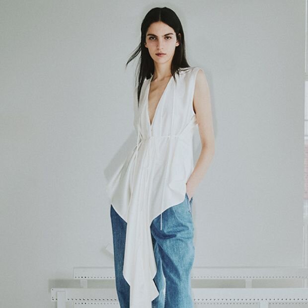 A model wearing a white sleeveless silk top with exaggerated scarf detail and silk tie closure.