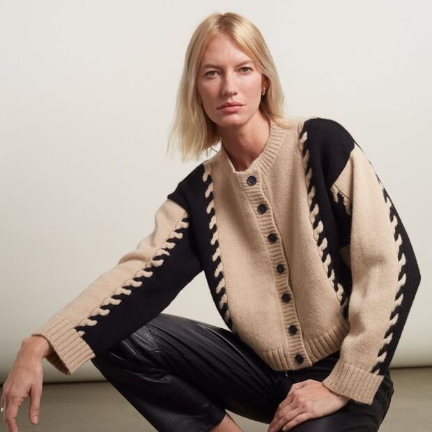 A model wearing a black and cream color block cable knit woll cardigan with dropped shoulders and a slightly cropped hem.