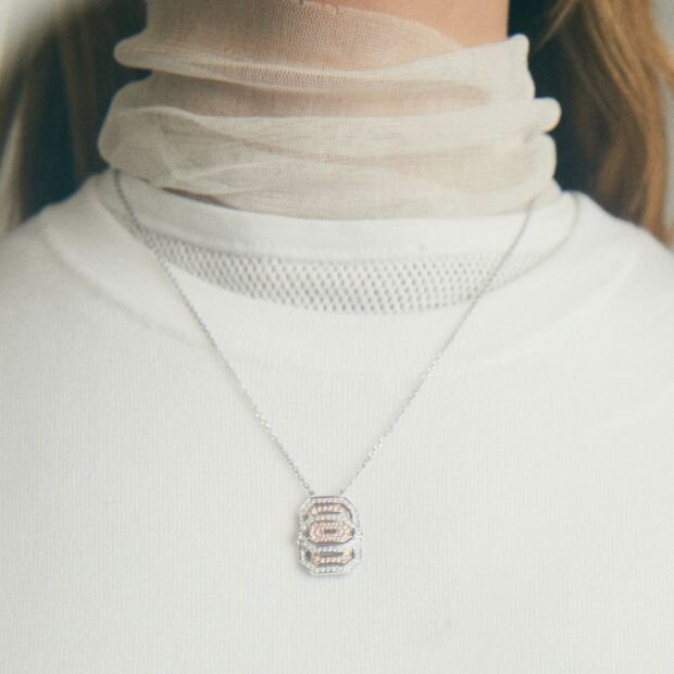 A person wearing a silver and rose gold diamond-studded pendant necklace over a white sheer turtleneck sweater.