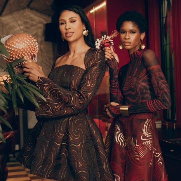 A model wearing a patterned jacquard mini dress with a domed neckline, flared sleeves, and a cinched waist; and another model wearing a patterned red and black mesh mock neck bodysuit with matching wide leg pants.