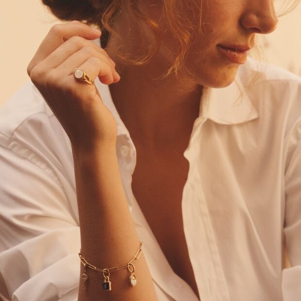 A woman wearing a gold ring with circular stone and a gold bracelet featuring multiple dangling charms.