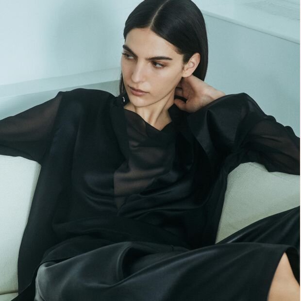 A model wearing a satin black midi dress with a black sheer long sleeve shirt.