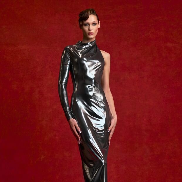 A model wearing a silver metallic asymmetric gown  with a single long sleeve and diagonally cutout back.