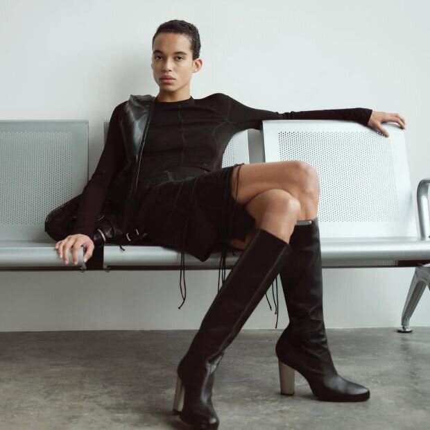 A woman wearing a long sleeve black top and a knit skirt with a garter-cut hem, and finishing off the look with black knee high boots.