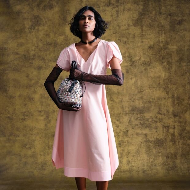 A model wearing a power pink poplin midi dress, features subtly layered cap sleeves, a V neckline, and ruching along the waist for a cinched silhouette.