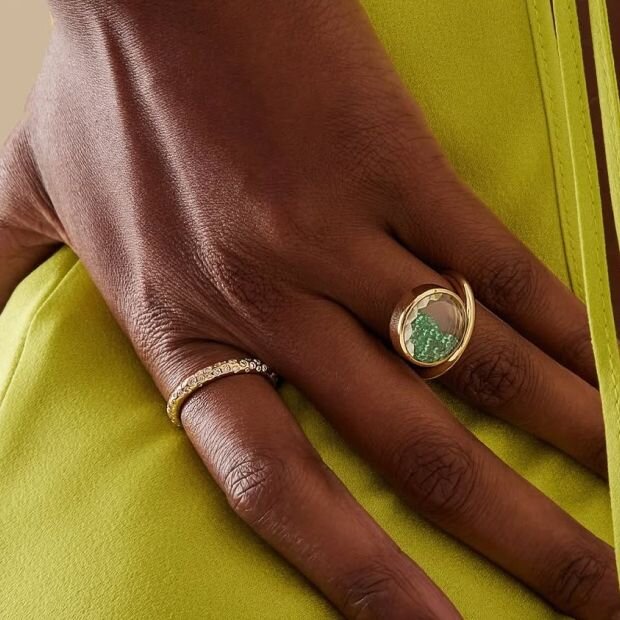 A woman wearing a golf ring with emeralds along and within a transparent sapphire crystal case.