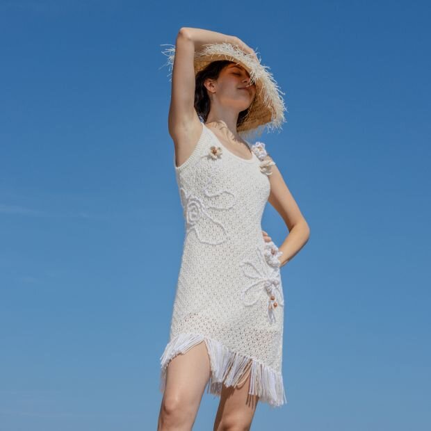 A model in a straw hat wearing a white dress with openworked crochet construction, featuring beaded florals and fringe at the hem.